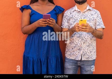 Anonymous multiracial couple standing messaging on mobile phones on background of orange wall in city Stock Photo