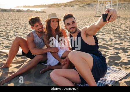 Young men wearing shorts and shirts and long haired woman in straw hat sitting on striped plaid on spacious sandy beach talking selfie Stock Photo
