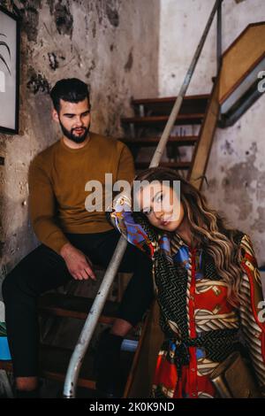 young fashionable woman looking at camera near young man sitting on a staircase inside a rustic mediterranean house Stock Photo