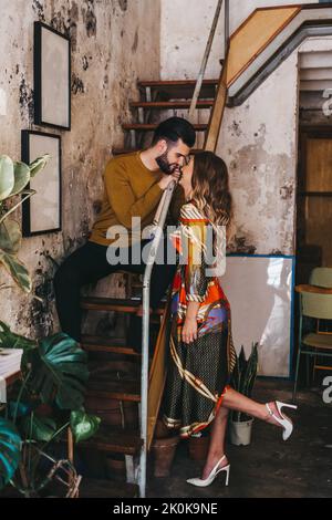 young fashionable woman looking kissing a young man sitting on a staircase inside a rustic mediterranean house Stock Photo