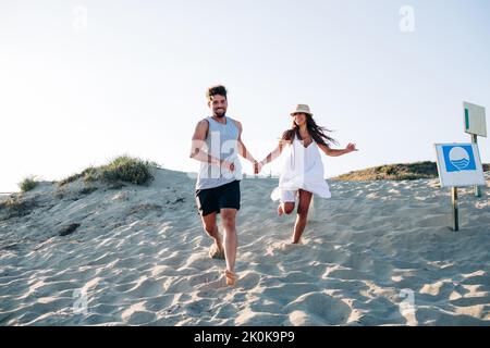 Glad barefoot couple in love wearing summer clothes running along sunlit sandy beach holding hands and toothy smiling Stock Photo
