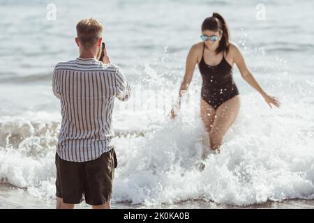 Couple on the beach while the man takes pictures of his wife with his mobile on a sunny day Stock Photo
