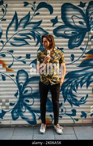 Full body of trendy young male with long hair holding cold ice cream in one hand and keeping another in pocket while standing against painted wall and Stock Photo