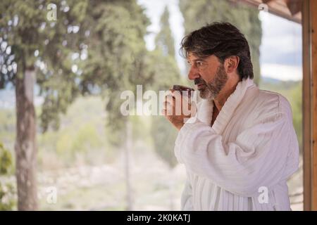 Contemplated senior gray haired male dressed in white soft bathrobe standing with cup of tea at terrace and looking away Stock Photo