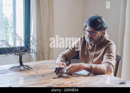 Side view of focused aged bearded male writing in notebook while sitting alone at home at daytime Stock Photo
