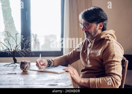 Side view of focused aged bearded male writing in notebook while sitting alone at home at daytime Stock Photo