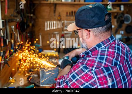 Side view of mature male worker using angle grinder while working with metal detail with glowing sparks in garage Stock Photo