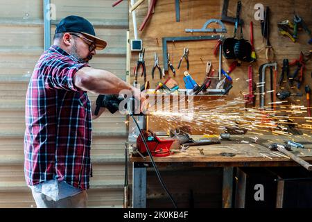 Side view of mature male worker using angle grinder while working with metal detail with glowing sparks in garage Stock Photo