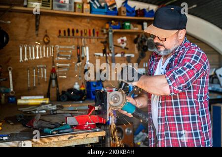 Side view of mature male worker using angle grinder while working with metal detail with glowing sparks in garage Stock Photo