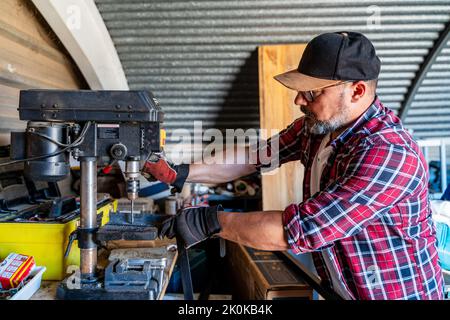 Side view of middle aged man in cap and plaid shirt working with wooden detail on drilling machine in light garage Stock Photo