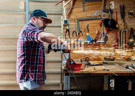 Side view of mature male worker using angle grinder while working with metal detail with glowing sparks in garage Stock Photo