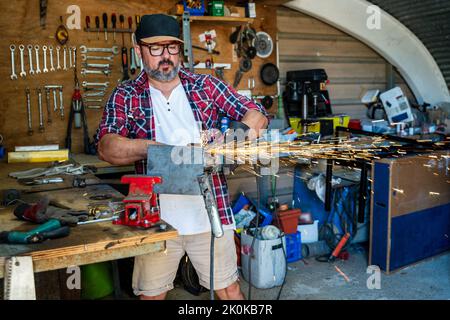 Mature male worker using angle grinder while working with metal detail with glowing sparks in garage Stock Photo