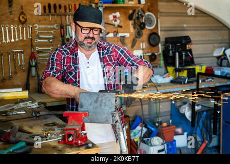 Mature male worker using angle grinder while working with metal detail with glowing sparks in garage Stock Photo