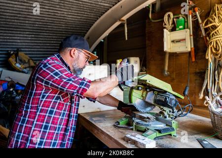 Focused mature man in cap and gloves working on miter machine while creating clean precise cut Stock Photo