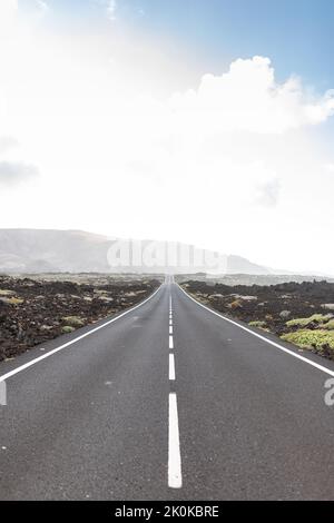 Long straight asphalt road running through volcanic terrain towards mountain against cloudy sky in Lanzarote Stock Photo