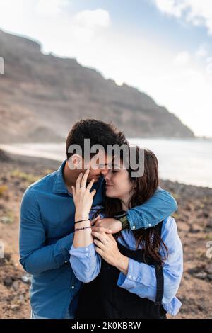 Side view of young ethnic guy in casual clothes embracing stylish girlfriend while relaxing together on sandy terrain with dry plants near mountains i Stock Photo