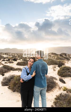 Back view of young ethnic guy in casual clothes embracing stylish girlfriend while relaxing together on sandy terrain with dry plants near mountains i Stock Photo