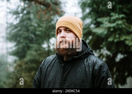 Thoughtful young man in coat with water drops and hat looking at camera in forest on blurred background Stock Photo