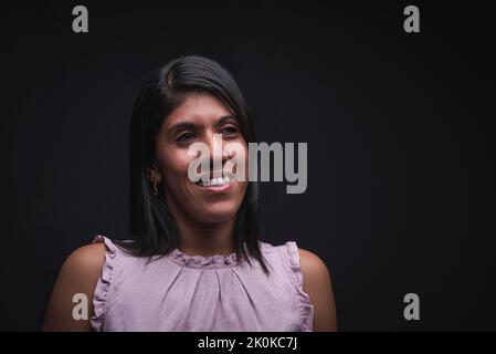 Positive self assured young Latino female wearing casual blouse smiling and looking away while standing against black background Stock Photo