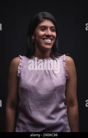 Positive self assured young Latino female wearing casual blouse smiling and looking away while standing against black background Stock Photo