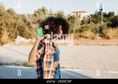 Side view of African American female hipster with afro hairstyle standing holding on the back a penny board on empty road and looking at camera during Stock Photo