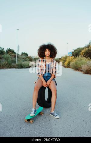 Happy black female with in trendy summer outfit and with afro hairstyle sitting on tire on an asphalt road with green penny board and looking at camer Stock Photo