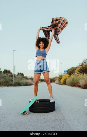 From below cheerful black female with in trendy summer outfit and with afro hairstyle sitting on tire on an asphalt road with green penny board and lo Stock Photo