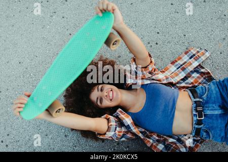 Happy black female with in trendy summer outfit and with afro hairstyle lying down on the asphalt road with green penny board and looking at camera Stock Photo