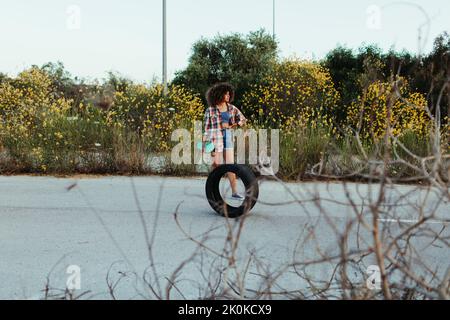 Happy black female with in trendy summer outfit and with afro hairstyle sitting on tire on an asphalt road with green penny board and looking away Stock Photo