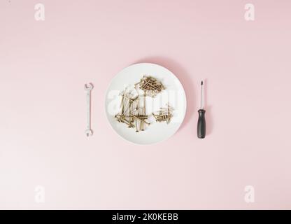 Screws served on a white plate with a screwdriver and a wrench on a pale pink background. Stock Photo
