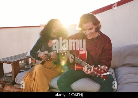 Excited young multiethnic friends in casual wear playing acoustic guitar and ukulele while sitting together on comfy couch on sunny rooftop Stock Photo