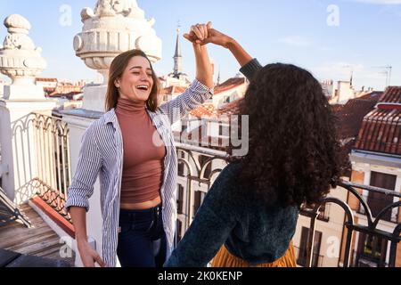 Excited young multiracial female friends wearing casual outfits holding hands and dancing together while spending sunny day on building rooftop Stock Photo