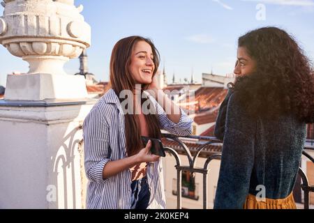 Content young multiethnic female friends in casual wear having conversation while standing together on sunny terrace in historic town district Stock Photo