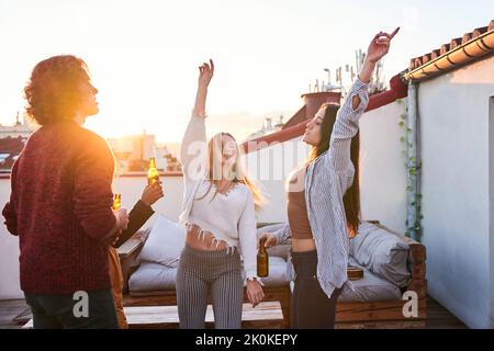 Group of joyful young multiracial friends wearing casual clothes enjoying cold beer and dancing with arms raised together on rooftop at sunset Stock Photo