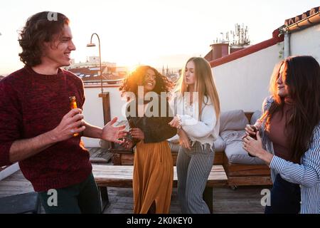 Group of happy young multiracial friends in casual clothes drinking cold beer and dancing together while having party on building rooftop at sunset Stock Photo