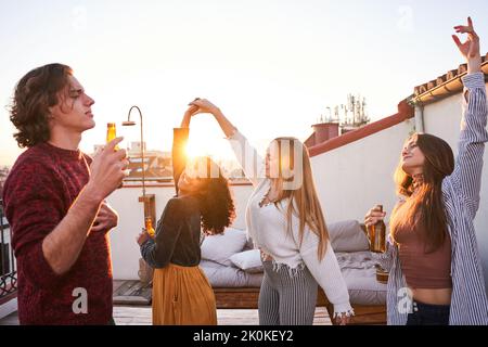 Group of happy young multiracial friends in casual clothes drinking cold beer and dancing together while having party on building rooftop at sunset Stock Photo