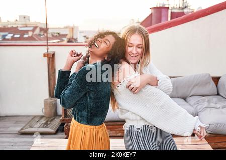 Cheerful young multiethnic female friends in casual outfits dancing and laughing merrily while spending warm day on building terrace Stock Photo