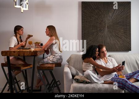 Smiling multiethnic couple in domestic clothes browsing mobile phone on comfy couch near female flatmates having dinner in living room Stock Photo