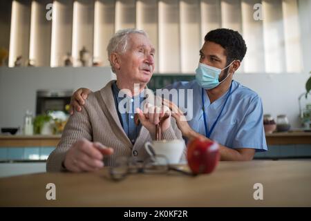 Caregiver with medical mask bringing healthy snack to senior man in nursing home care center. Stock Photo