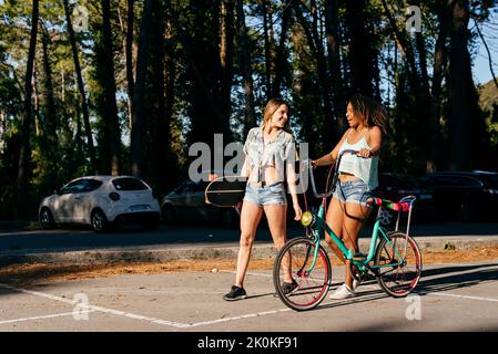 Female friends walking on street holding skate board and bicycle in vacations Stock Photo