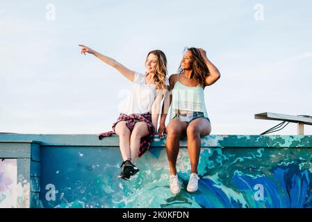 Two attractive multiethnic cheerful women sitting together on a colorful wall leaning on each other's shoulder while looking away Stock Photo
