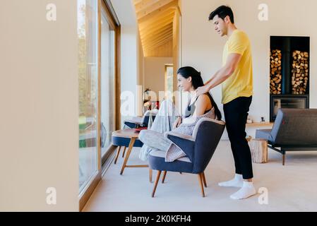 Young man giving a massage to girlfriend on chair in room and looking at big window Stock Photo