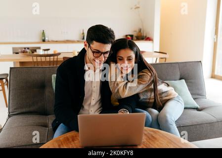 Young guy in eyeglasses typing on laptop near cheerful attractive Hispanic lady on settee Stock Photo