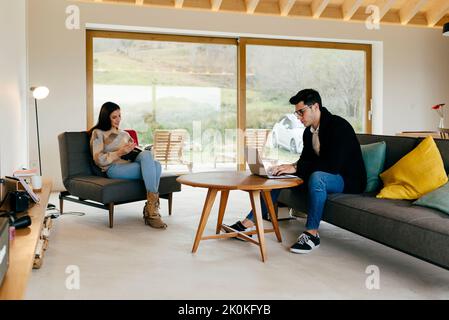 Young attractive couple on settee man at laptop on table and woman reading in room Stock Photo