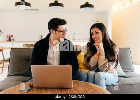 Young guy in eyeglasses typing on laptop near cheerful attractive Hispanic lady on settee Stock Photo