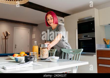 Young female with bright pink hair taking jar with cereals from table while cooking breakfast in morning in modern apartment Stock Photo