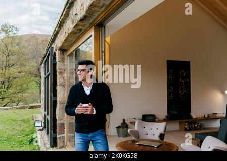 Young man in casual wear drinking from mug near modern cottage between hills Stock Photo