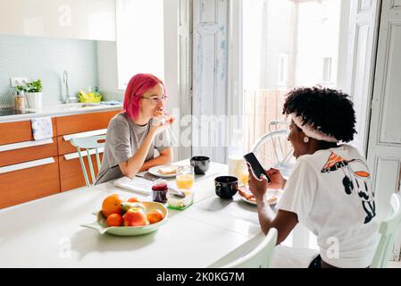 Young woman browsing smartphone while having breakfast at home with friend Stock Photo
