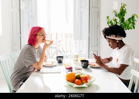 Young woman browsing smartphone while having breakfast at home with friend Stock Photo