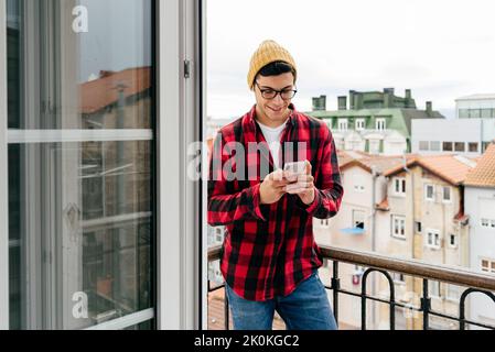 Attractive hipster caucasian man with beanie hat an glasses standing on balcony at home using mobile phone Stock Photo